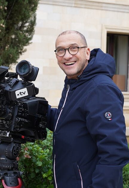 Smiling cameraman with a large TV camera in an outdoor setting near a building.