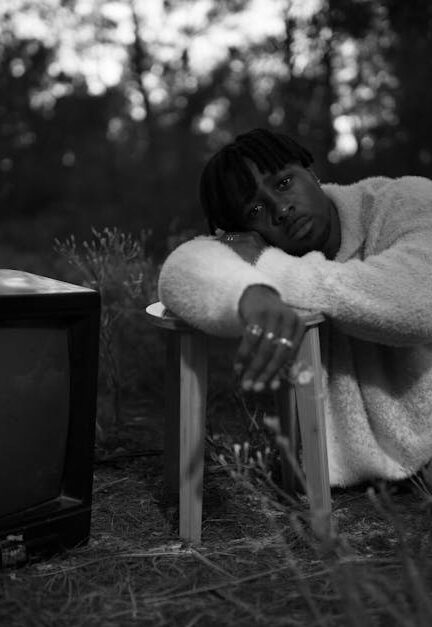 A man in a sweater leans on a stool next to a retro TV in a forest setting, evoking nostalgia.