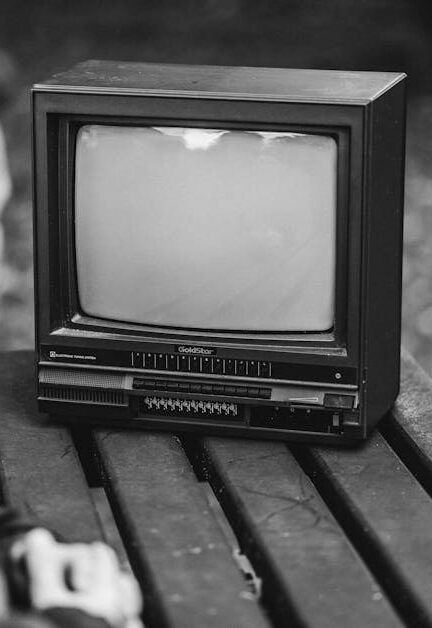 Black and white side view of anonymous male sitting at wooden table and looking at old fashioned television in nature on blurred background