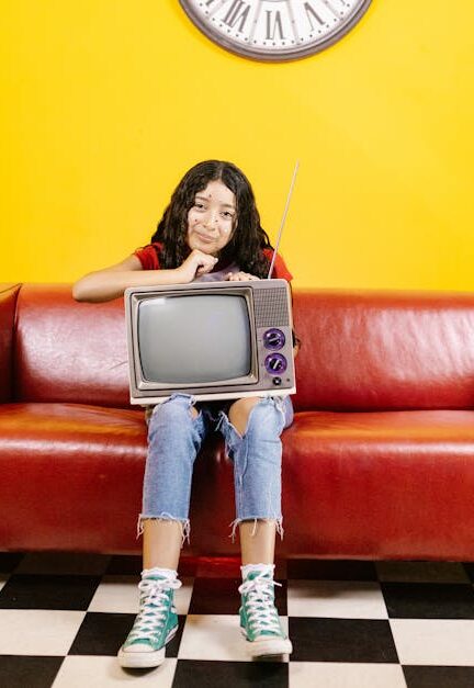 A young woman with a CRT TV sits on a red couch in a colorful living room.
