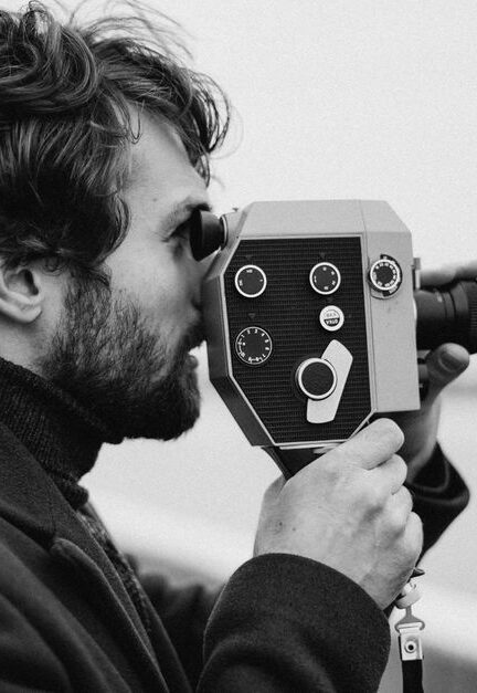 Side view of a man filming with a vintage camera in a black and white setting.
