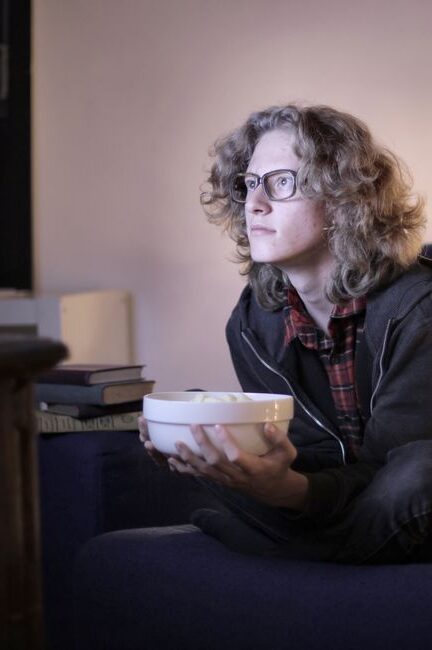 Concentrated male teenage with long hair in casual wear and eyeglasses sitting on couch with bowl of popcorn and watching interesting movie while resting in cozy living room at home
