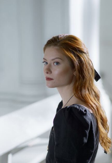 Portrait of a woman in a black dress sitting indoors, looking away thoughtfully.