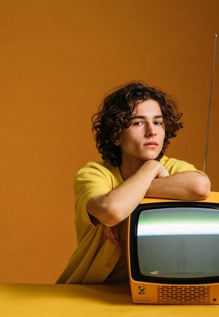 Trendy young man with curly hair in yellow fashion posing with an old TV set.