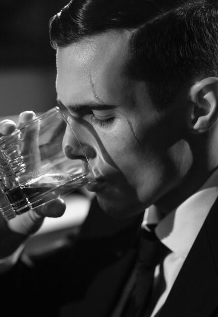 Black and white image of a man sipping whiskey, evoking classic film noir style.