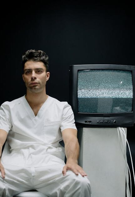 A serious man in white attire sits beside vintage TVs with static screens.
