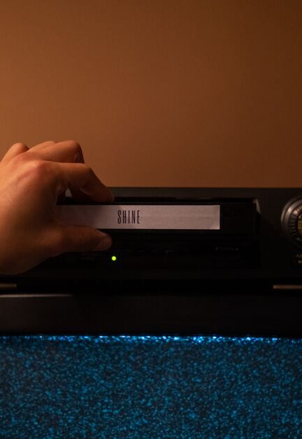 A hand inserting a VHS tape labeled 'SHINE' into a vintage player. Nostalgic home movie setup.